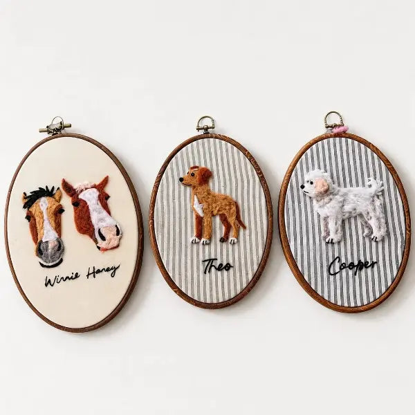A collection of three custom handmade 3D needle-felted pet portraits in oval wooden hoops, including two horse heads on cream fabric, a full-body brown dog on striped fabric, and a full-body fluffy white poodle on striped fabric.