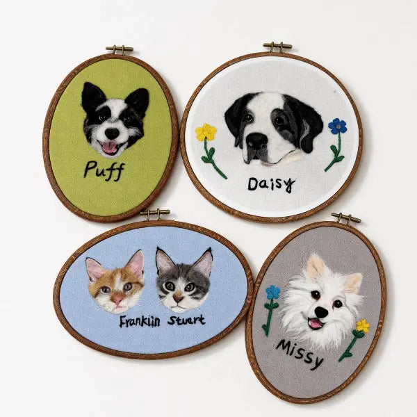 A collection of four custom handmade 3D needle-felted pet portraits in wooden embroidery hoops, including a black and white corgi on green fabric, a St. Bernard dog on white, two cats on blue, and a white fluffy Pomeranian on grey fabric.